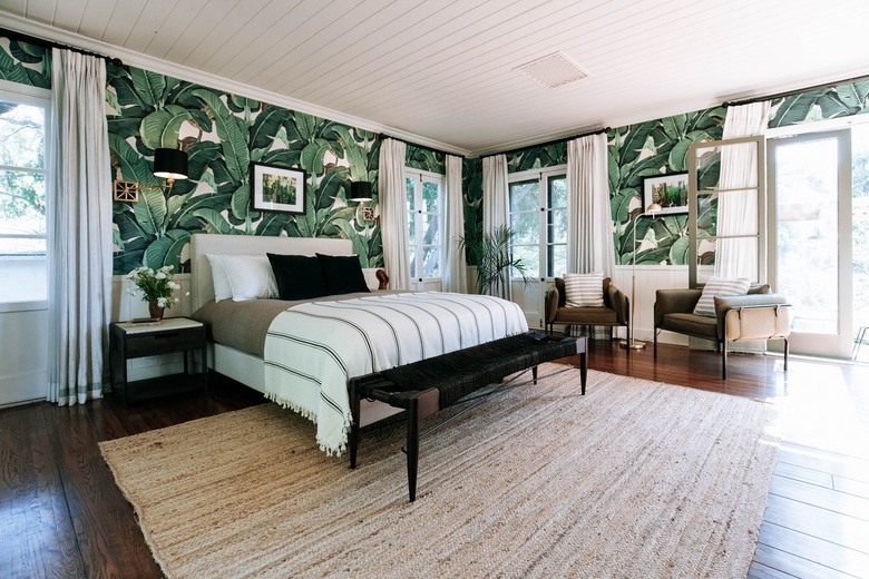 Bedroom with tropical palm wallpaper, French door windows, black-gray-white bedding, neutral fiber rug, and wood flooring