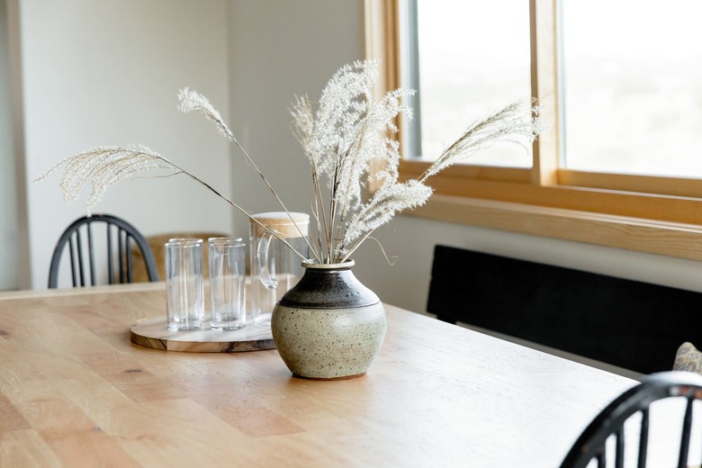 A light wood rectangular dining room table. On the table, there is a grey and black ceramic vase with stalks of wispy grass in front of a set of glasses and a glass pitcher. There are two black Windsor chairs at the table, as well as a black bench. The bench is under a large window.