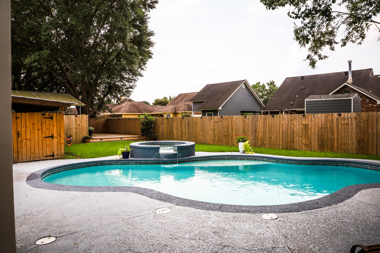 A large free form gray grey accent swimming pool with turquoise blue water in a fenced in backyard in a suburb neighborhood.