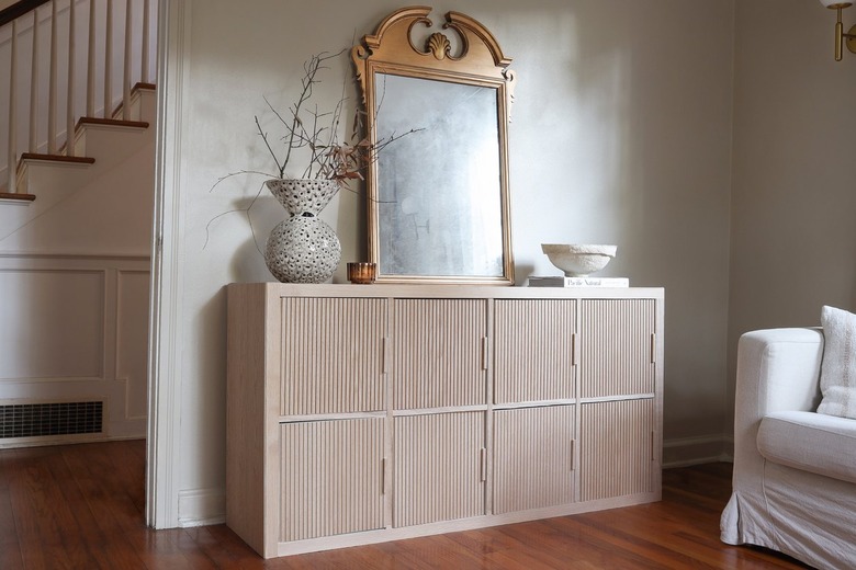 IKEA hack KALLAX shelf with fluted wood doors styled with a gold mirror, vase, and candle