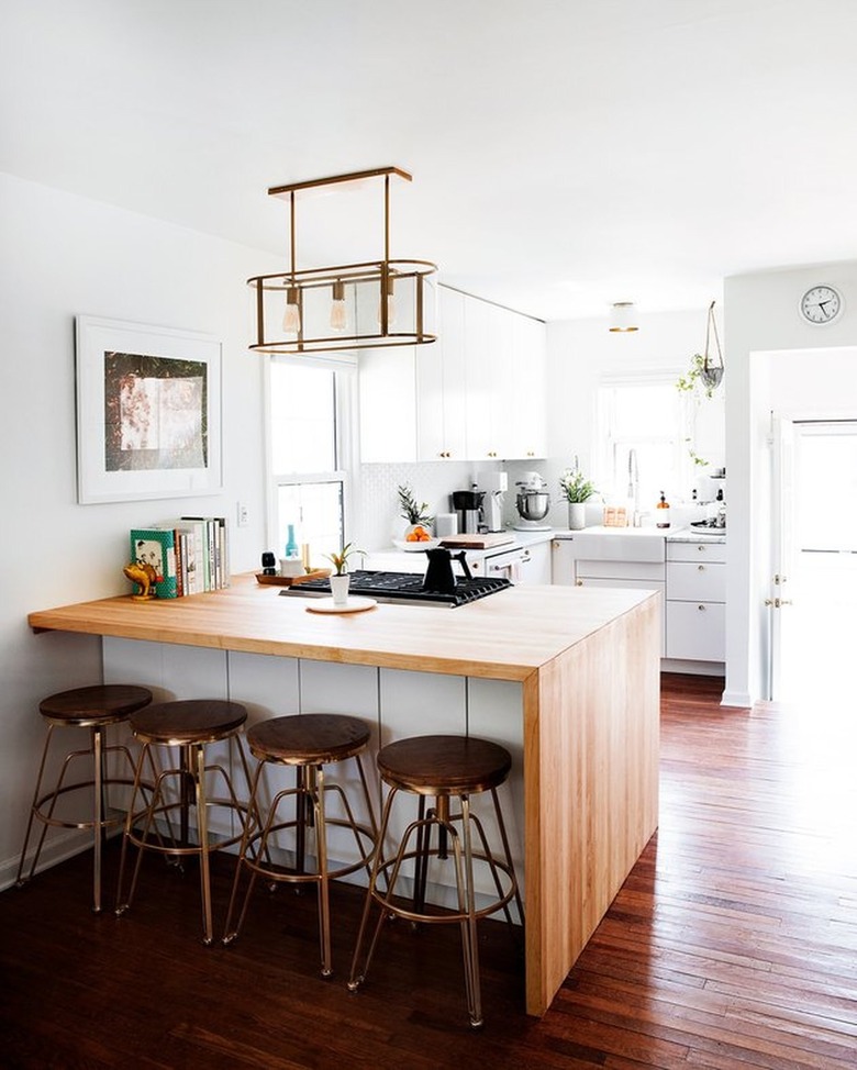 Small kitchen with wood waterfall island, bar stools, white cabinets, wood floor, pendant light.