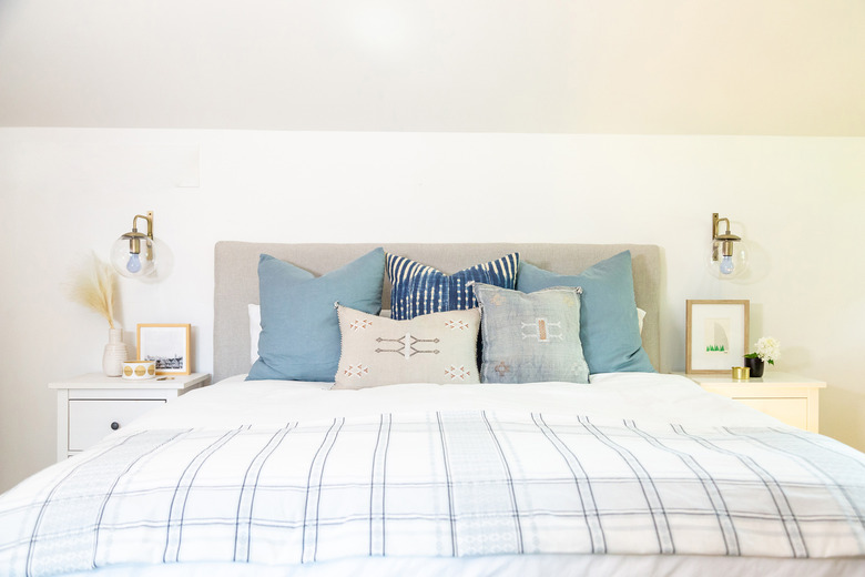 Upholstered headboard with brass sconces and many pillows on bed.