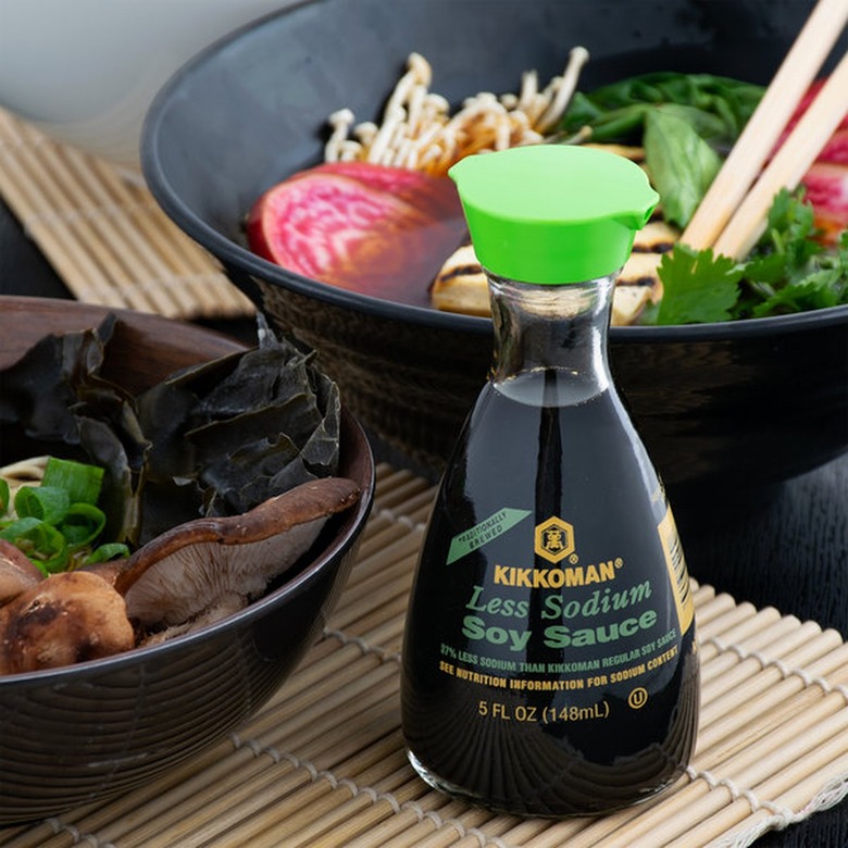 A clear glass bottle of soy sauce with a bright green top on a bamboo mat on a table with black bowls of food.