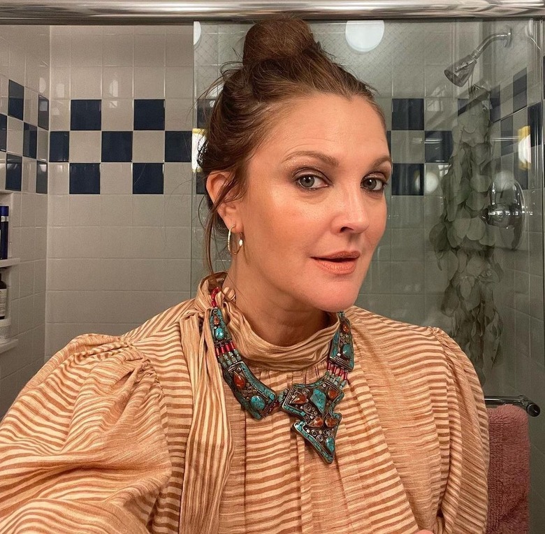 A photo of actress Drew Barrymore in front of her glass shower door with her hair up in a bun, a turquoise chunky necklace, and a brown and white striped top.