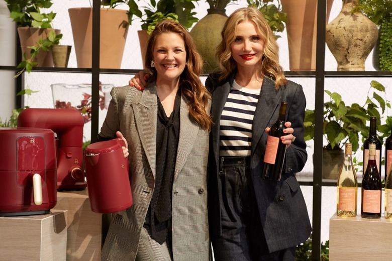 Drew Barrymore and Cameron Diaz stand in front of a wall of plants with their arms over each others shoulders. Barrymore holds her Merlot electric kettle from her brand Beautiful by Drew, while Diaz holds a bottle of Merlot from her brand, Avaline. Barrymore is wearing a light grey, oversized blazer with a black top underneath, and Diaz is wearing a black blazer with a white and black striped top. To Diaz's side is more wine from her label, and to Barrymore's side is a Merlot colored air fryer and stand mixer.