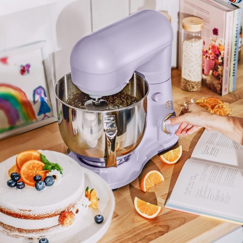 A lavender stand mixer on a wooden countertop next to a white cake topped with fruit.