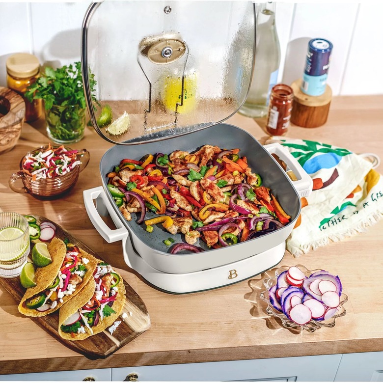 A light blue and white electric skillet full of vegetables on a wooden countertop next to a board topped with tacos.