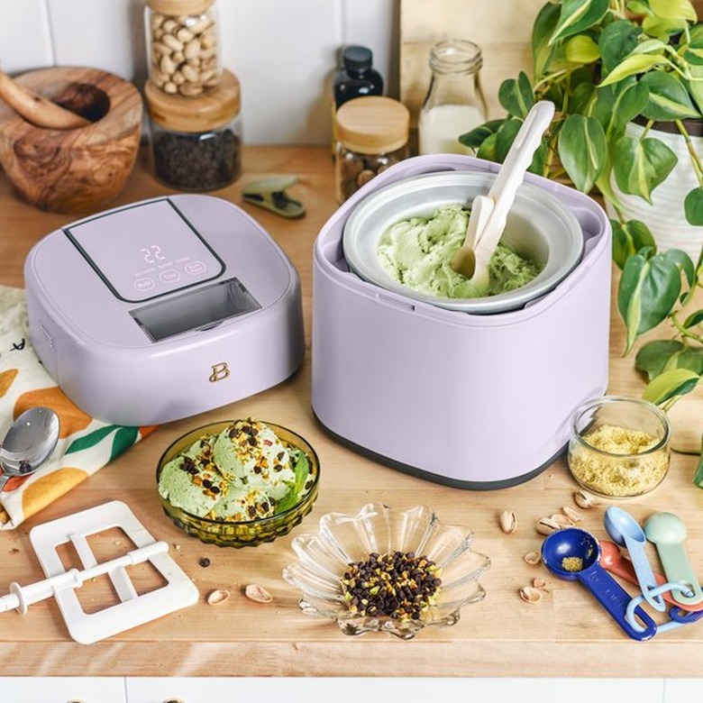 A lavender ice cream maker full of green ice cream next to some bowls and measuring spoons on a wooden counter.