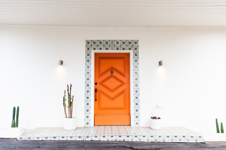 An orange front entrance door with geometric accents. Blue floral tile decorate the frame and landing. Cacti plants are on either side.