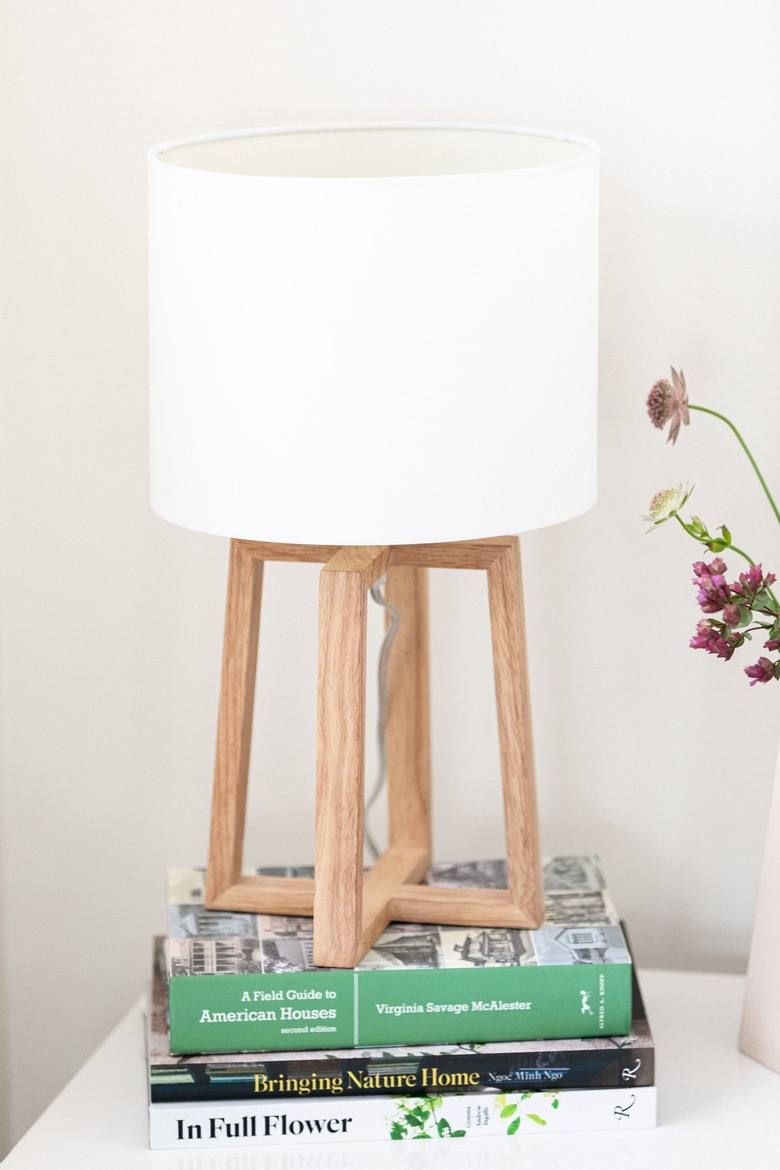 A lamp with a white shade and a wood base on top of books on a nightstand