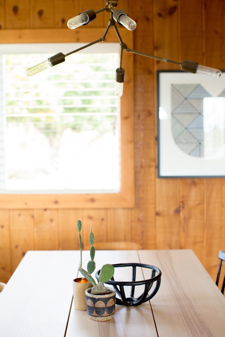 Wood dining table, open black bowl and cacti in planters, in a wood walled dining room with a Modern multi-bulb chandelier.