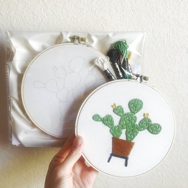 person holding embroidery hoop with cactus pattern near kit with another hoop and embroidery and linen