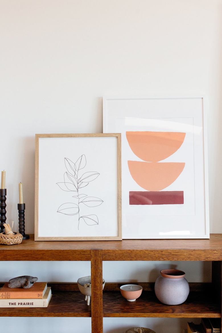 Two framed pieces of art next to two candles in candlestick holders on top of a wooden bookcase. On the shelf below, a small stack of books and ceramic pots and candle holders.