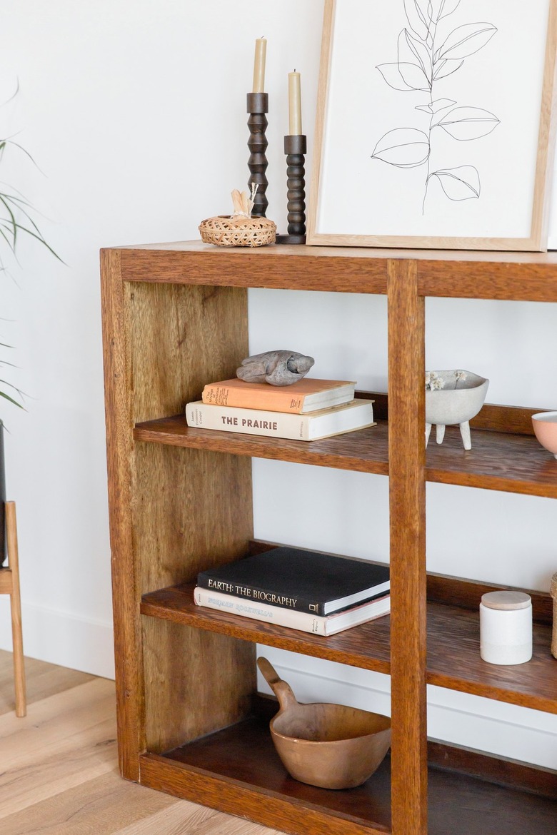 Detail of a wooden bookshelf with books and other decorative trinkets such as ceramic pots and a woven basket. Two candlesticks in black holders and a framed drawing of leaves on top.