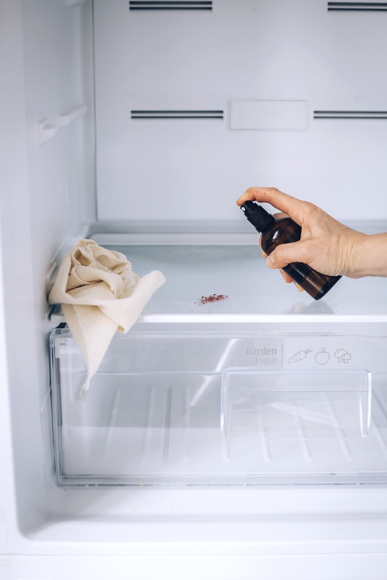 Hand using spray bottle and paper towels to clean inside shelf of white fridge