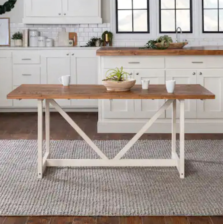 Wood farmhouse table with white trestle base, rug, kitchen cabinets.