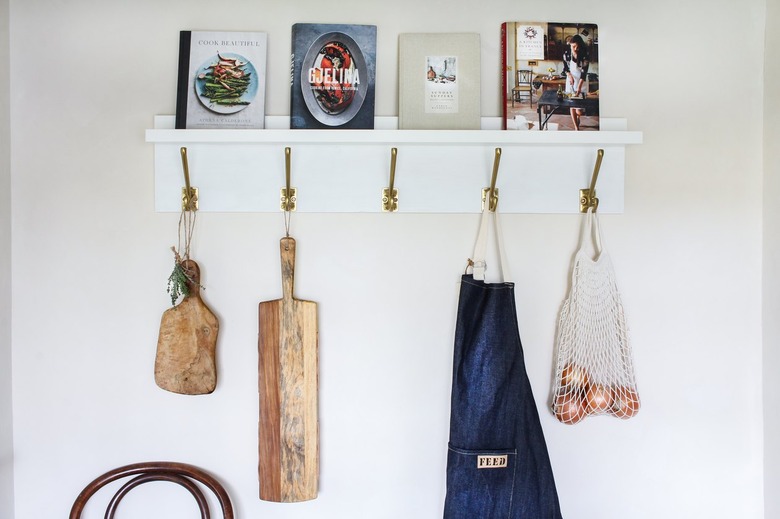 White kitchen wall hangers with gold hooks holding cutting boards, blue apron, and bag of tomatoes on white wall with books resting on top