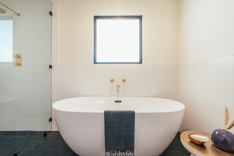 white freestanding tub with gray towel draped over, gold wall-mounted faucet and handles, white wall tile on half of the bathroom wall, small table with bathroom accessories, gray floor tile, glass shower door