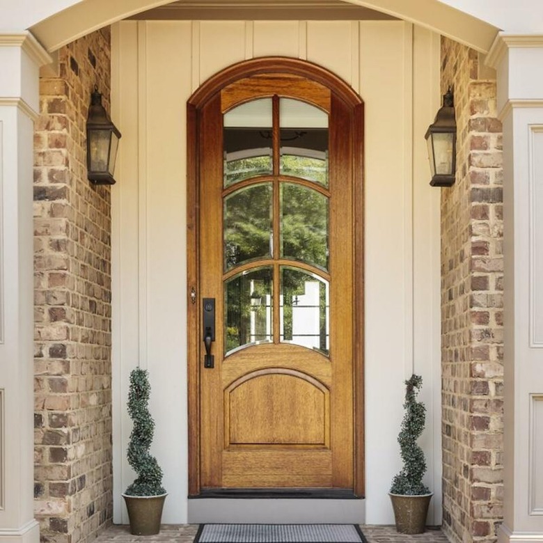 A wooden arched front door on a brick house; two matching planters with evergreens are on either side of the door