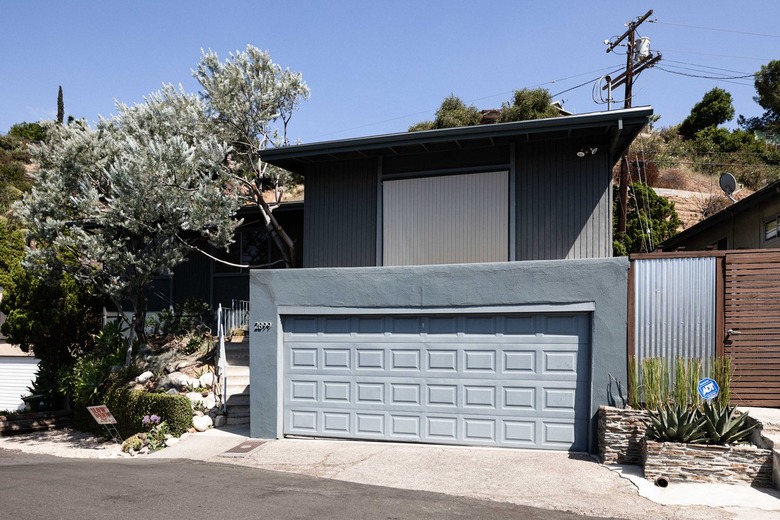 Gray house with a large garage, wood and corrugated metal fence, stone planters, and trees.