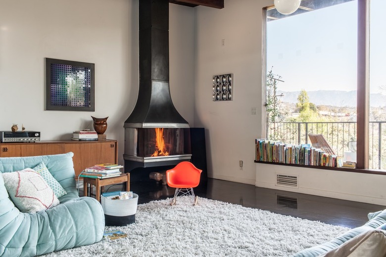 A blue sofa bed on a shag carpet. A small orange rocking chair. Wood furniture and a stove fireplace. Row of books by picture windows.