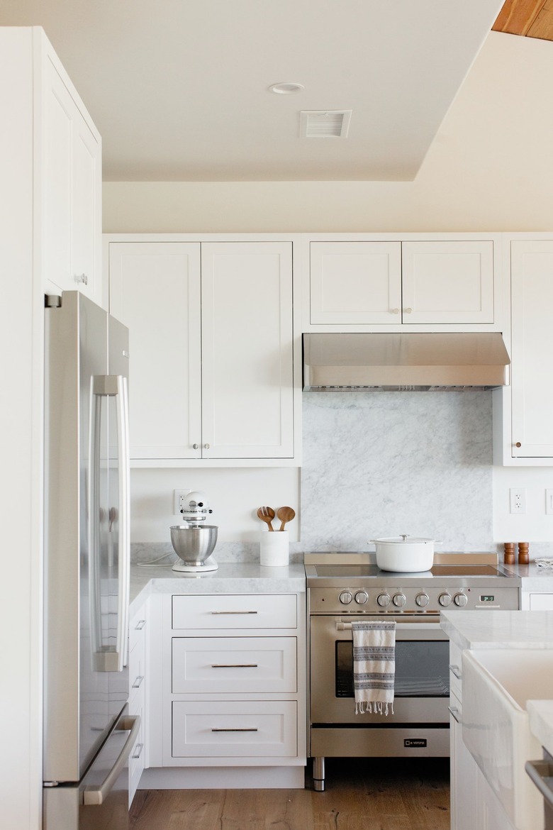 Kitchen with a stainless steel range, white cabinets, and a marble countertop. On the countertop, there's a white KitchenAid stand mixer with a stainless steel mixing bowl along with a white utensil holder with wooden salad tongs. On the stovetop, there's a white dutch oven. To the left, there's a stainless steel refrigerator. To the right, there's a white kitchen island with a sink.