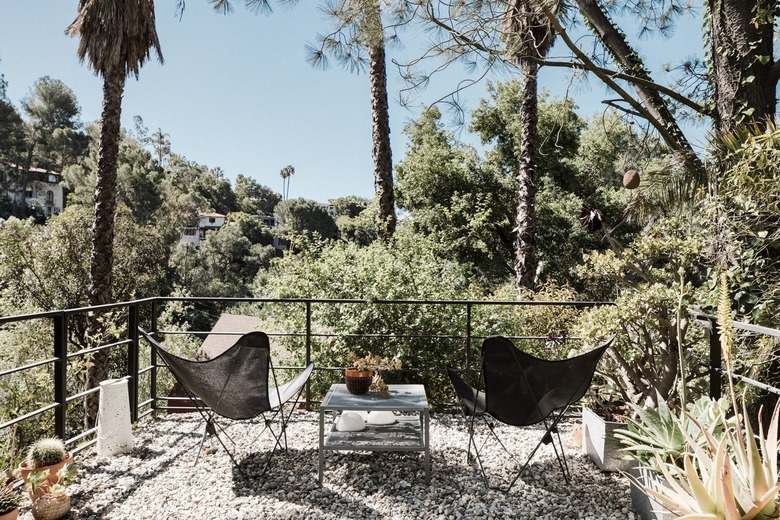 Gray butterfly chairs, square table on balcony with black railing and gravel. Palm trees surround.