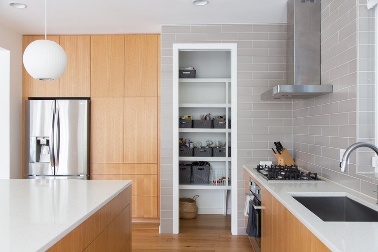 Kitchen with wood cabinets, round white pendant light, white counters, gray tiles, wood floors. A pantry with gray baskets.