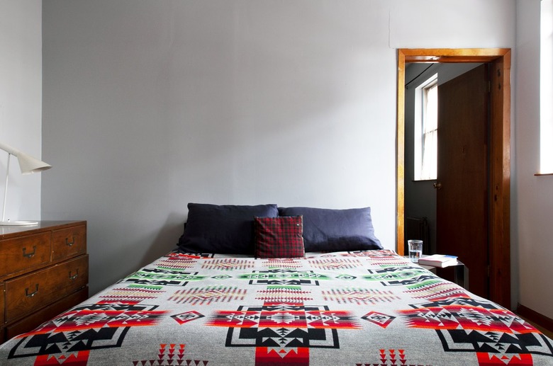 Bed with gray pillows, red plaid accent pillow, Southwest bedspread, and a wood dresser with a white lamp.