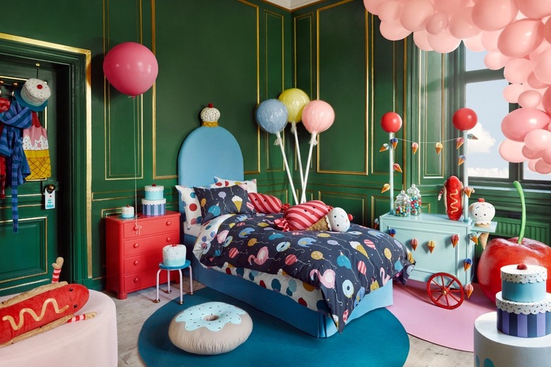 A child's bedroom. There are green walls, and a pink balloon arch around a window. The bed at the center of the room is blue, with a navy blue comforter with flamingos, balloons and ice cream cones on it. There is a play food cart by the window, with a stuffed hot dog and ice cream cone on it, draped with an ice cream cone garland. A giant faux cherry sits next to it, with play cakes on a table by it as well. A blue striped robe, a cake-shaped hat, and an apron that looks like an ice cream cone hangs on the door.