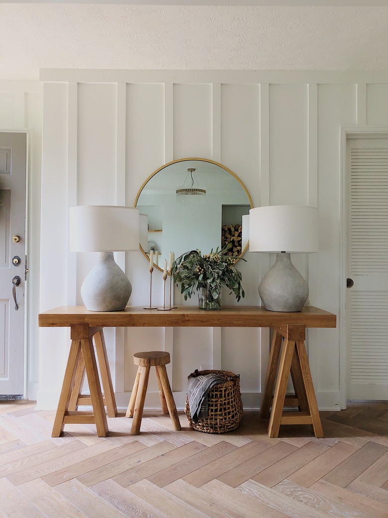 Entryway with rustic table, matching lamps, round mirror, wood floor, basket, wood stool, plant.