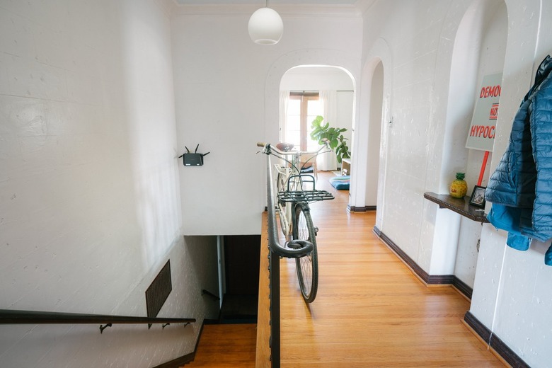 A wood staircase with a black banister. A hallway with arched wall niches, with shelves. A bicycle and a blue jacket.