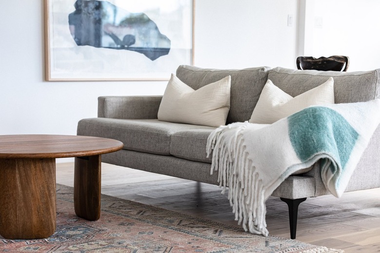 Grey couch with white pillows and white tasseled blanket in contemporary living room with round wood coffee table, framed art, and rug