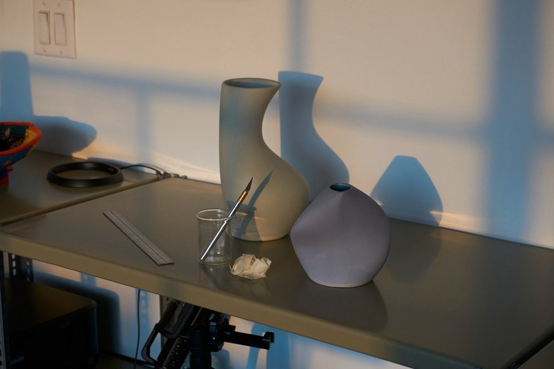 A tall green curvy vase and short blue curvy case on a metal table next to a glass cup with a paintbrush in it.