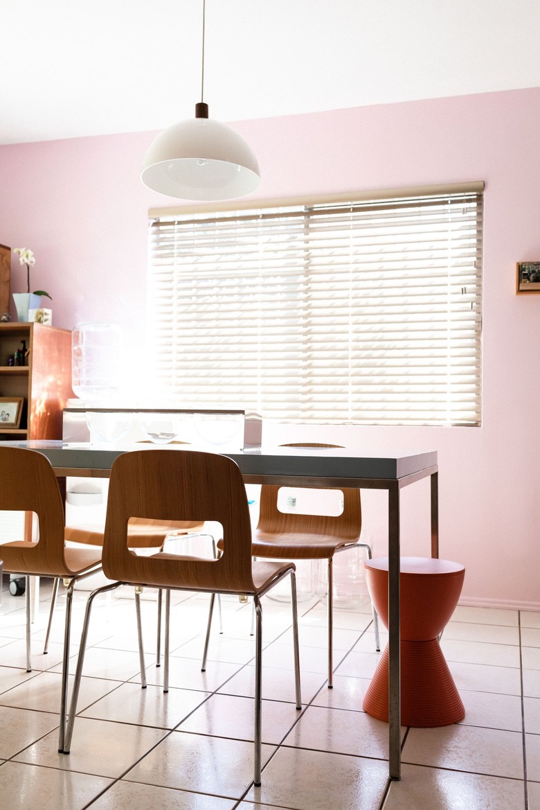 Dining room with pink wall, white tile floor, wood chairs, gray table, orange drum stool, and white bell pendant light.