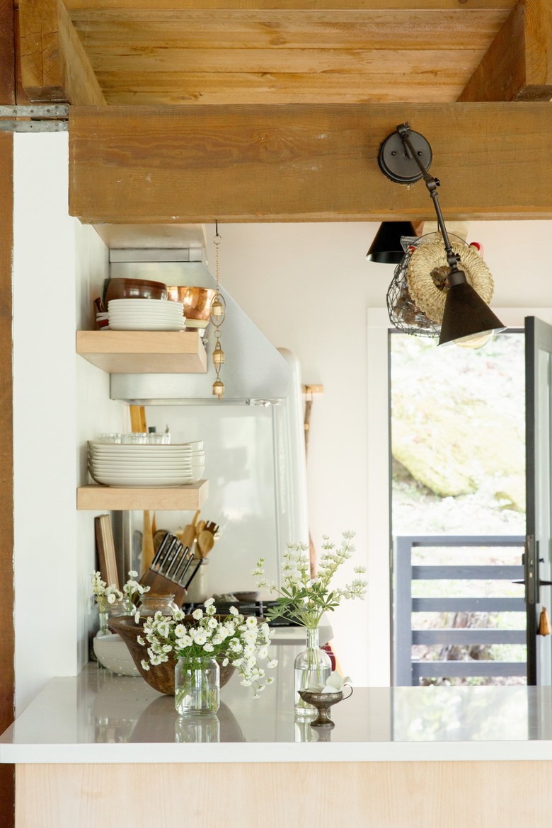 Kitchen with light wood shelves and a white counter. Decorated with flowers in clear vases, and a black lamp sconce. A wood ceiling.