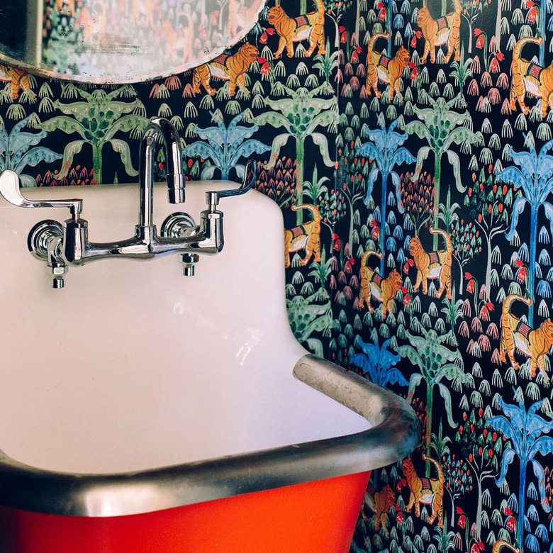 A maximalist tiger and palm tree wallpaper next to a red bathroom sink with silver fixtures and a white interior.