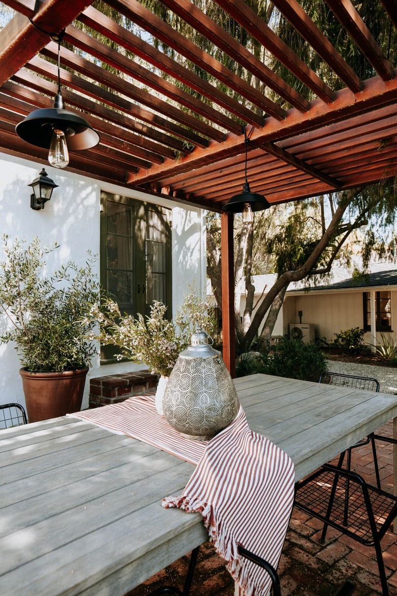 Wood pergola over a wood outdoor dining table with black wire chairs on a brick patio