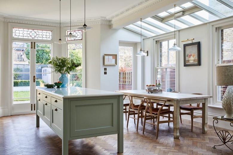 A kitchen with white walls, crown molding, a light green marble kitchen island, and a large dining table under a skylighted ceiling.