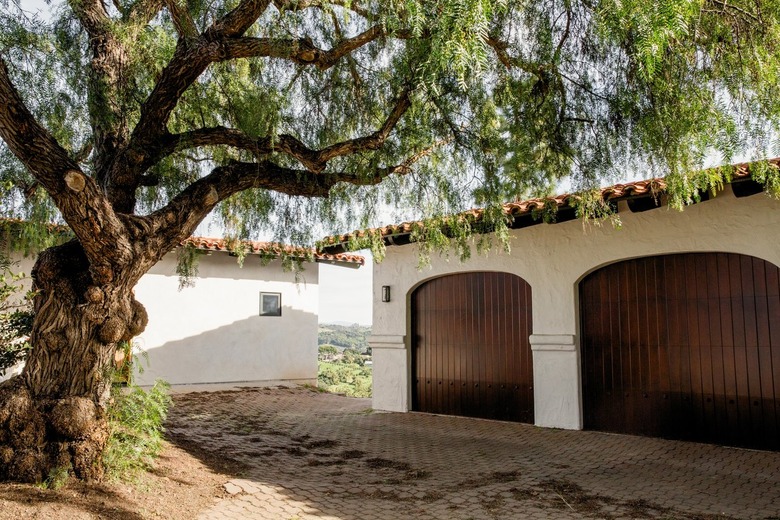 Brick driveway leading to a white Spanish-style garage with two sleek wooden doors. A large oak tree grows on the other side of the driveway, its branches reaching towards the garage Home Exterior Ideas