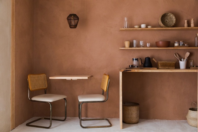 Woven pendant light over a square dining table and cane chairs. Brown walls and wood shelves with eclectic dish ware, and a wood table.