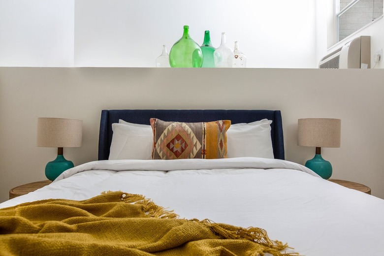 Modern eclectic bedroom with dark upholstered headboard, matching green lamps, gold blanket, southwestern pillow.