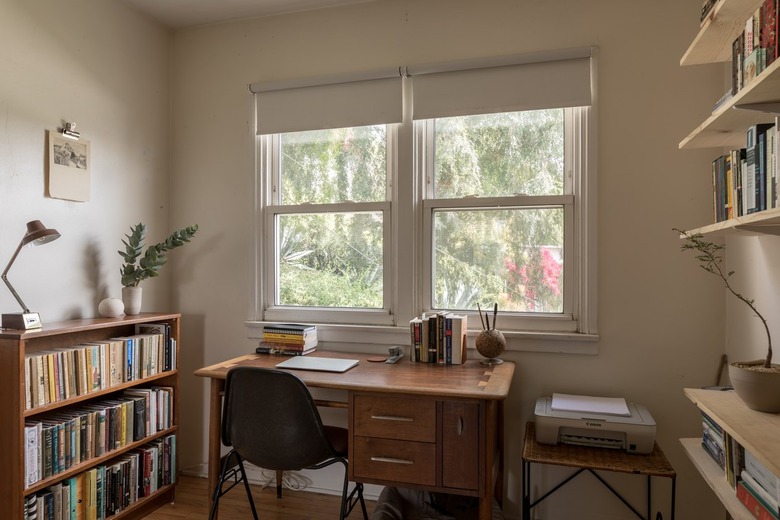 Wood office desk surrounded by bookshelves and shelves of books. Houseplants decorate the space. A printer is on a small table.