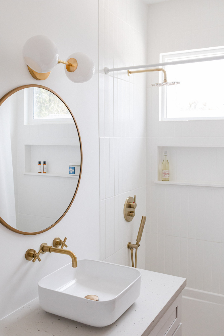 white ceramic vessel sink, gold faucet, round mirror with gold trim, white and round light fixture, white tiled shower with gold fixtures