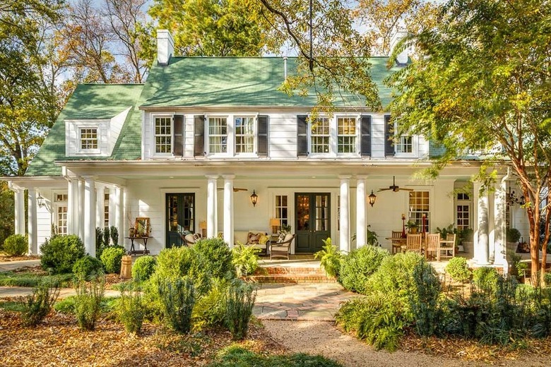 A traditional white house with a green roof and black shutters has a fully landscaped yard and sitting porch.