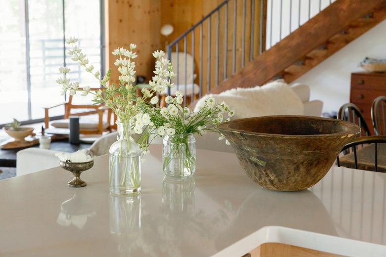 A white counter with vases of flowers, a wood bowl, and a small pewter dish. A wood staircase in the distance.