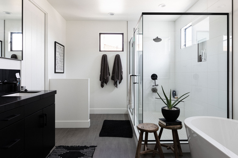 large bathroom with freestanding white tub, two wooden stools, black vanity, glass shower with black trim, two towels hung up, wood flooring