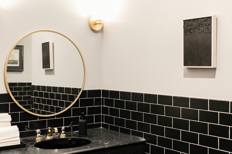 black subway tile wall, black vanity with gold fixtures, gold round mirror, rectangular artwork with black interior