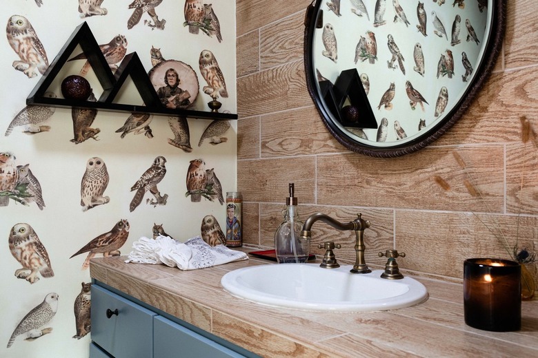 A wood bathroom countertop with round mirror, wood-like subway tile backsplash, and wallpaper with owls