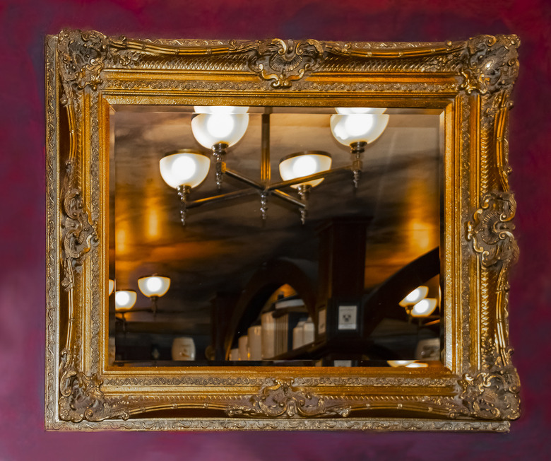 A dark red faux painted marbilized wall with an ornate golden mirror reflecting a warm rustic room with light fixture and shelving